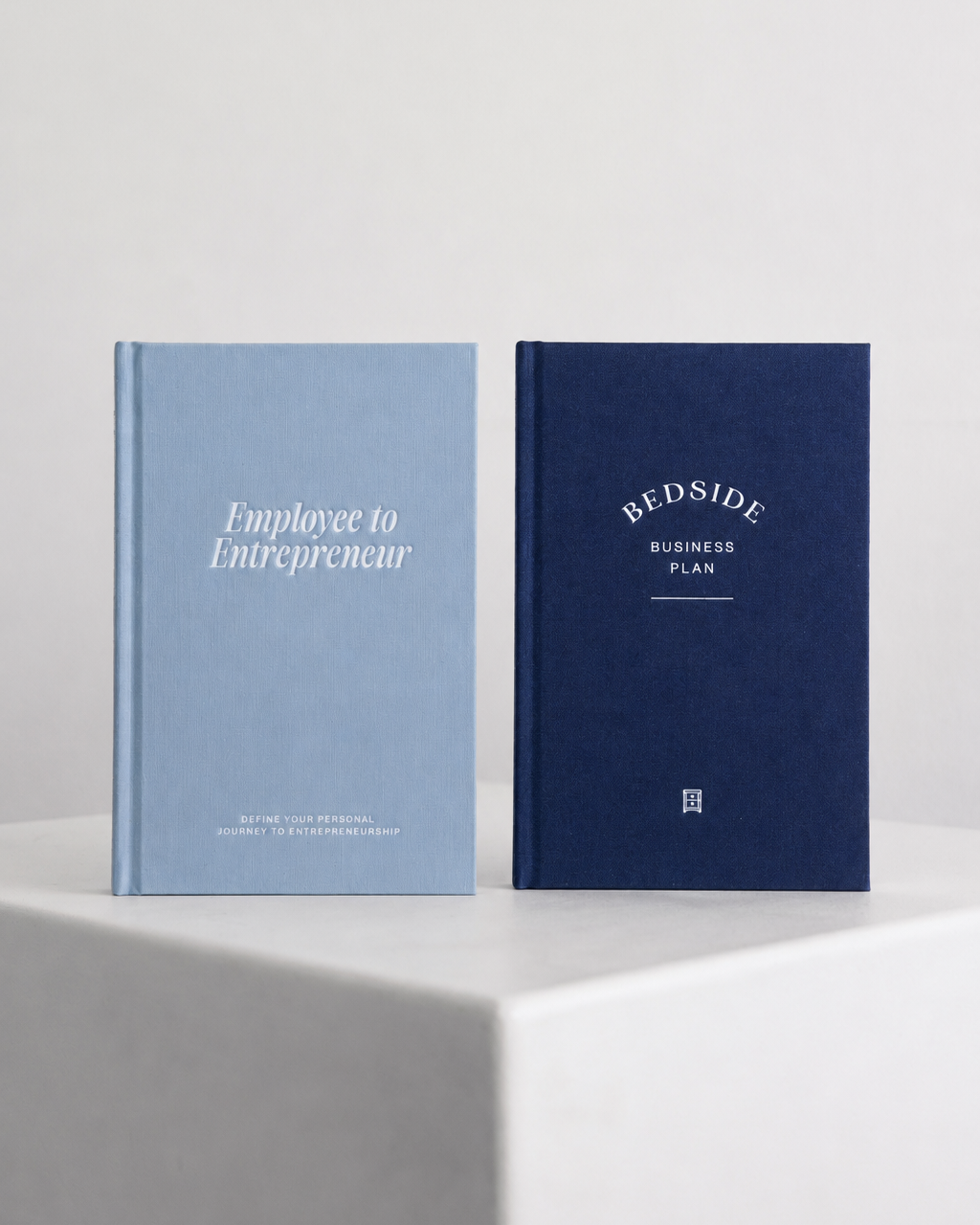 Two hardcover books standing upright on a white pedestal against a light gray wall. The left book is light blue with white text reading 'Employee to Entrepreneur,' and the right book is dark navy with white text reading BEDSIDE BUSINESS PLAN